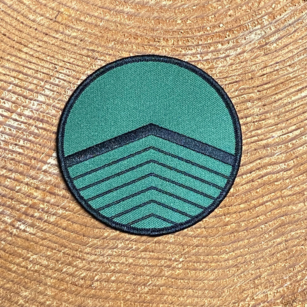 A round, dark green patch with black stitching on the edge and a pattern of clack chevrons facing down starting on the bottom half of the design.