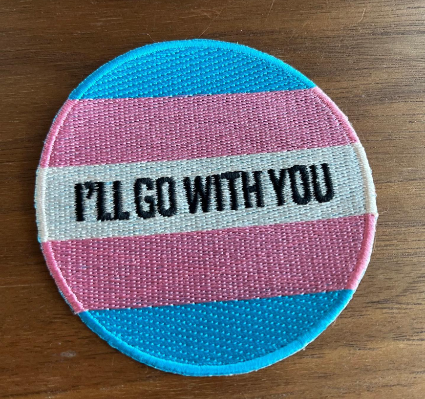 Round 3x3 embroidered patch with "I'LL GO WITH YOU" In the middle in black letters on a Trans flag background.